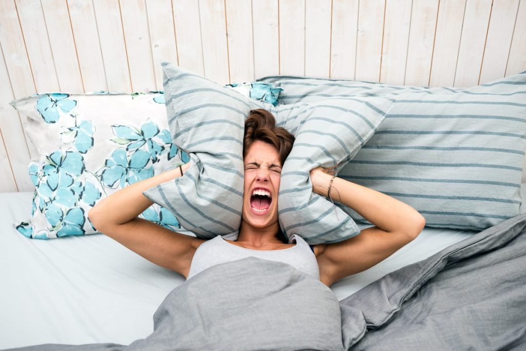 A lady in bed, screaming while holding head between her pillow, out of frustration because she can't sleep.