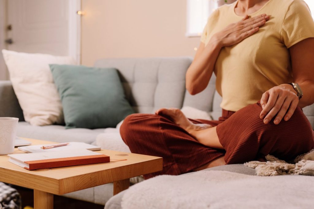 A woman sitting on the couch, holding her heart center. Breathing deeply with a journal next to her, destressing and regulating the nervous system.