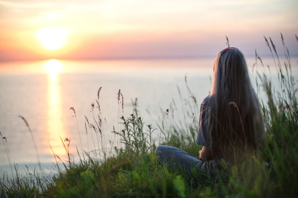 A woman sitting in the grass looking out at the sunset on the lake, finding peace.