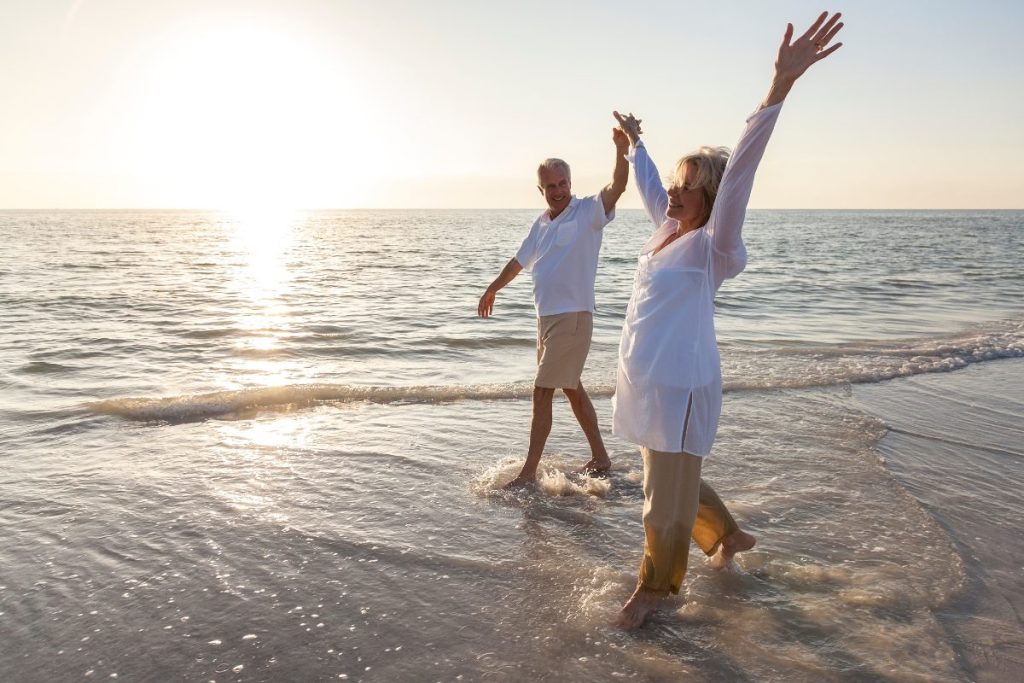 Husband and wife smiling happily walking barefoot on a beach.