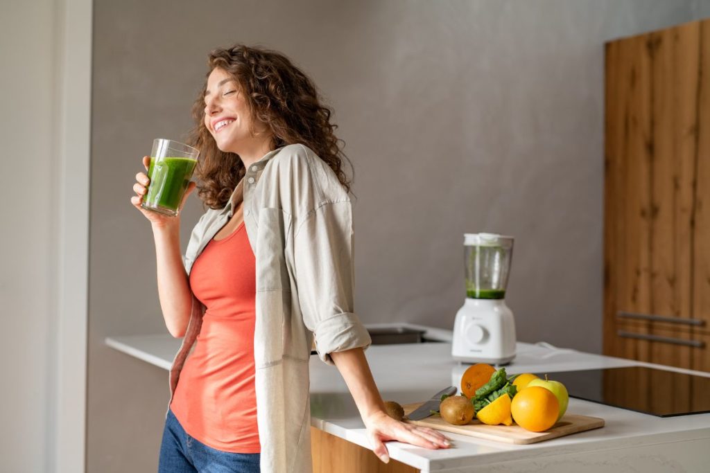 A woman smiling, drinking a green juice next to her juicer.